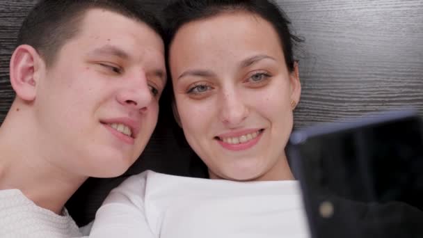A young couple holding a smartphone in hand is photographed together, shoots video blog on bed. Happy family husband and wife take photos online using a modern mobile phone at home on sofa in bedroom. — Stock Video