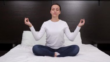 Nilüfer pozisyonunda meditasyon yapan rahat genç bir kadın evde yatakta oturuyor. Evde kapalı gözlerle yoga antrenmanı, ellerle meditasyondan zevk almak. Akşam evde meditasyon yogası yapan bir kadın.