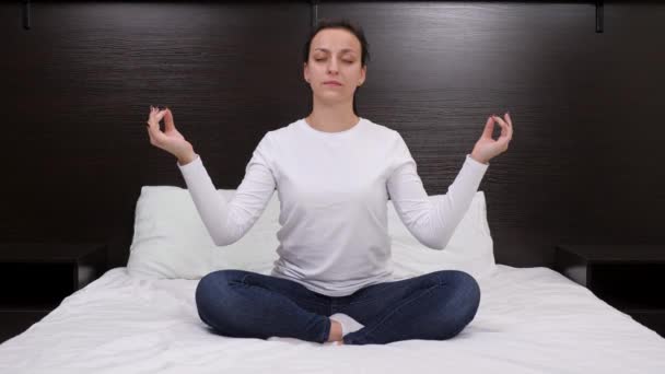 Mujer joven y relajada meditando en posición de loto sentada en la cama en casa. Practica yoga con los ojos cerrados en casa, disfrutando de la meditación con las manos. Mujer relajante con meditación yoga por la noche en casa — Vídeo de stock