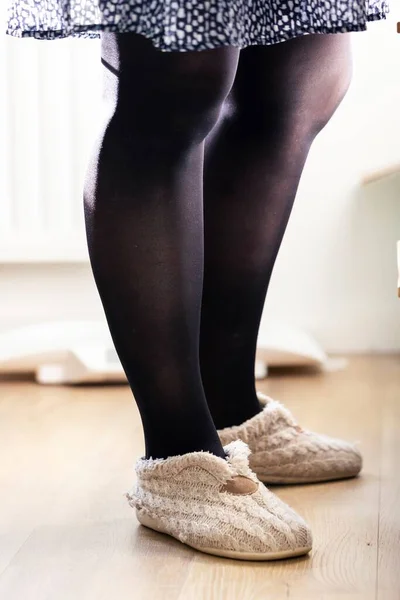 A portrait of female legs covered in black nylon pantyhose or stockings while wearing slippers or mules on the feet standing indoors on a wooden floor in front of a cupboard.