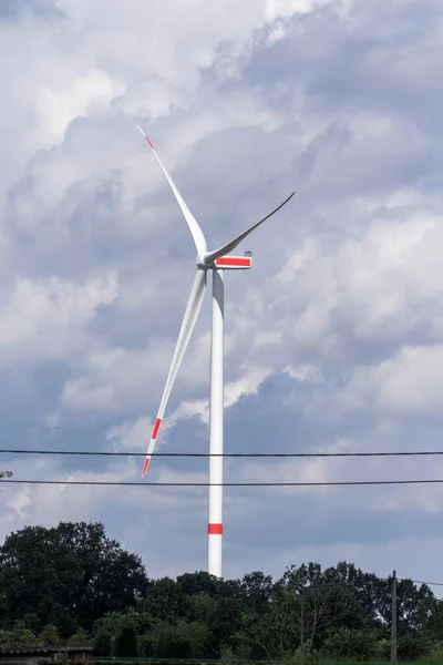 A portrait of a big windmill standing tall above the trees with a cloudy sky behind it. The turbine generates clean ecological sustainable and green electric energy by catching wind on its wings.