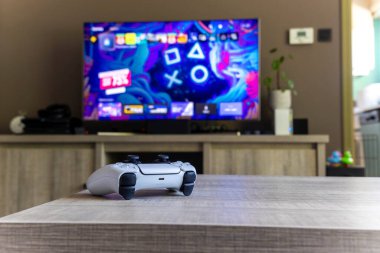 BRECHT, BELGIUM  AUGUST 14 2022 A portrait of a Sony Playstation 5 white basic wireless controller on a wooden table in front of a television showing the PS5 playstation plus screen in a living room