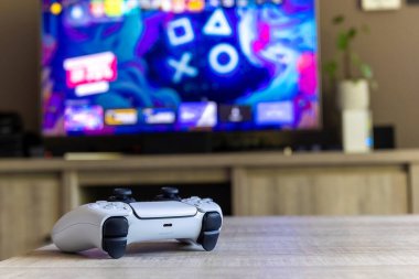 BRECHT, BELGIUM  AUGUST 14 2022 A portrait of a Sony Playstation 5 white basic wireless controller on a wooden table in front of a television showing the PS5 playstation plus screen in a living room