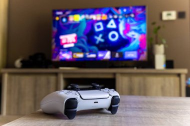BRECHT, BELGIUM  AUGUST 14 2022 A wide portrait of a Sony Playstation 5 controller on a wooden table in front of a television showing the PS5 playstation plus screen in a living room.