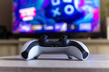 BRECHT, BELGIUM  AUGUST 14 2022 A portrait of an official Sony Playstation 5 wireless controller on a wooden table in front of a television showing the PS5 playstation plus screen in a living room.