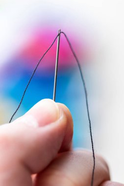 A portait of two fingers holding a metal sewing needle with a thread running through the eye of the needle. The wire is running through the hole and there is a colorful background.