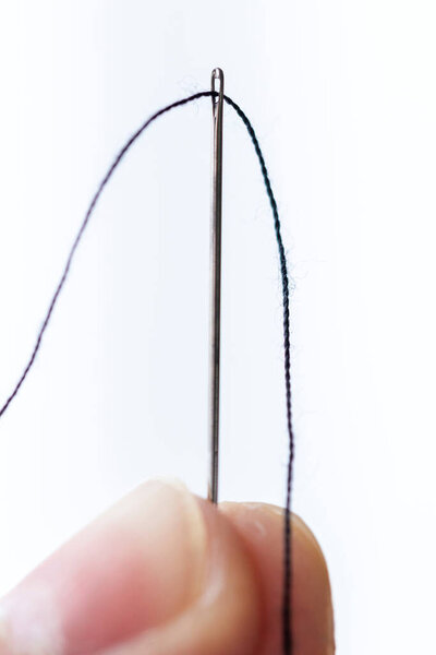 A portait of two fingers holding a metal sewing needle with a thread running through the eye of the needle on a white background. The wire is running through the hole.