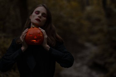 Genç, sıska bir kadının portresi. Ormanda parlayan bir balkabağı tutuyor.