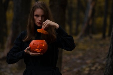 Genç, sıska bir kadının portresi. Ormanda parlayan bir balkabağı tutuyor.