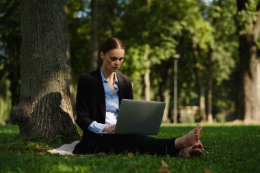 Yeşil çimlerin üzerinde oturan bir kadının yan görüntüsü. Elinde defter, dışarıda çalışan, uzaktan kumandalı bir kadın..