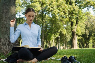 Yeşil çimenlerin üzerinde dizüstü bilgisayarlı, dışarıda çalışan, uzaktan kumandalı mutlu bir kadının yan görüntüsü..