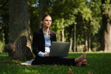 Yeşil çimlerin üzerinde oturan bir kadının yan görüntüsü. Elinde defter, dışarıda çalışan, uzaktan kumandalı bir kadın..