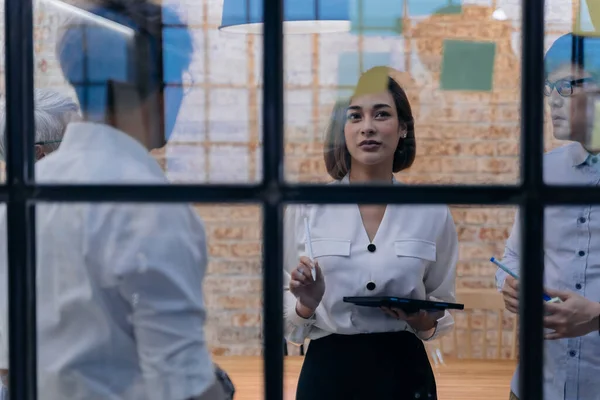 20s Asian company manager femenino lluvia de ideas en la sala de reuniones mientras tiene notas adhesivas en el vidrio en la oficina — Foto de Stock