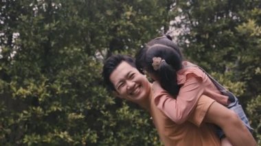 Genç bir baba kızını sırtında taşıyor ve parkta gülümsüyor.