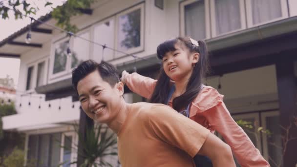 Ung asiatisk far ger sin dotter piggyback och ler framför huset — Stockvideo