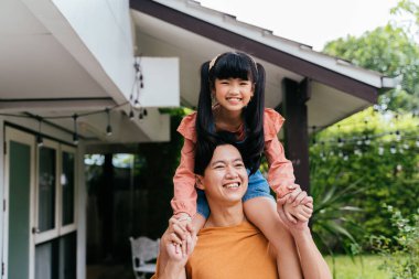 Genç baba, kızını sırtında taşıyor ve evin önünde gülümsüyor. Mutlu Asyalı ebeveynler