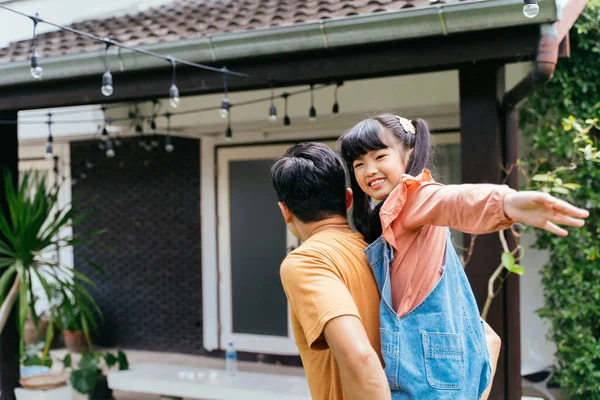 Joven padre dando a su hija a cuestas y sonriendo delante de la casa. feliz asiático padre. —  Fotos de Stock