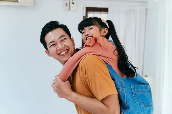Joven padre dando a su hija a cuestas y sonriendo delante de la casa. Feliz crianza asiática —  Fotos de Stock