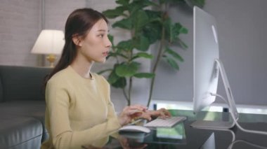 Genç Asyalı iş kadını evde çalışıyor ve oturma odasında masaüstü bilgisayarını kullanıyor.