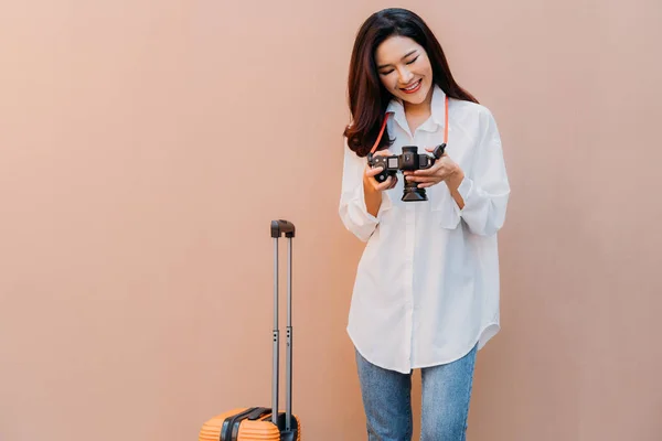 Young solo traveling Asian woman checking the photos from her vacation on camera with desert sand colored wall in background and orange suitcase next to her in casual clothing with copy space