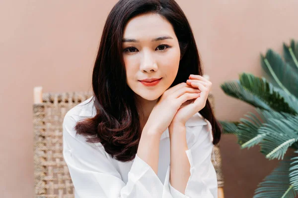 Portrait of a charismatic Asian female sitting on chair in white shirt with smiles, Young woman with red lips with a nice desert sand colored wall and palm leaves in background