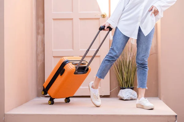 Young solo traveling woman standing with Georgian door as she is towing her bright orange suitcase filled with remote working tech, putting away plane tickets in her left pocket