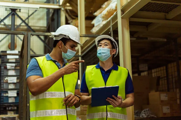Group of young factory warehouse workers wearing a protective face mask while working in logistic industry indoor. Asian and Indian ethnic men checking item order during Coronavirus Covid 19 pandemic