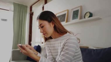30 'lu yaşlarda cep telefonu çalan Asyalı bir kadın evdeki oturma odasında koltukta oturuyor. Mutlu kadın internet ve teknoloji kullanıcısı rahat bir odada kanepede günlük giysiler içinde - 4K Slow Motion Video