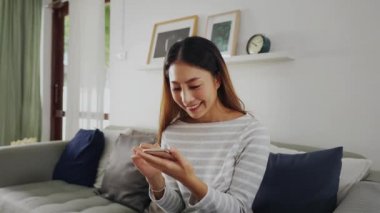 30 'lu yaşlarda cep telefonu çalan Asyalı bir kadın evdeki oturma odasında koltukta oturuyor. Mutlu kadın internet ve teknoloji kullanıcısı rahat bir odada kanepede günlük giysiler içinde - 4K Slow Motion Video
