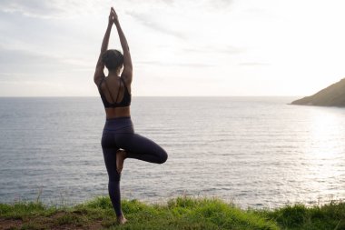 Genç ve güzel bir kadın, okyanus sahilinin kenarında, fotokopi aletiyle yoga yapıyor.