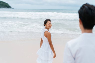 Mutlu gülümseyen 30 'lu yaşlardaki Asyalı kadın arkasını dönüyor ve yaz aylarında kumlu sahilden bir adam tarafından aranıyor. Beyaz gömlekli ve elbiseli çift - Romantik ilişki ve birlikte seyahat kavramı