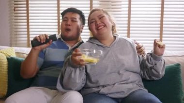 Young Asian chubby couple singing and watching tv on the couch. Man and woman enjoying a fun time together at home - 4K Slow motion Footage