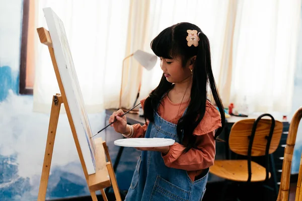 Cute Asian girl in casual clothes with pigtails holding palette and painting picture on canvas while standing near easel in light room at home