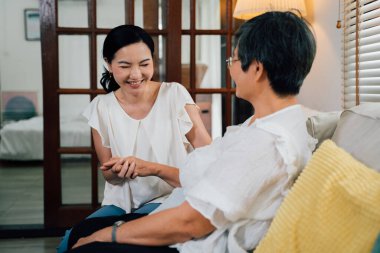 Neşeli, yetişkin Asyalı kız, gülümseyen ve yaşlı anneyle el ele tutuşan. Hafta sonu evde, oturma odasında kanepede otururken.