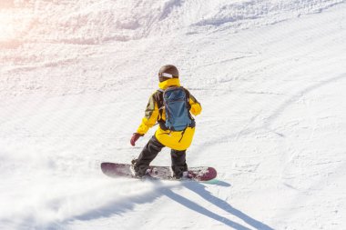 Kasklı bir snowboardcu ve sırtında bir sırt çantasıyla güneşli bir günde dağ yolunda geziyor.