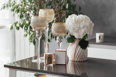Yüksek bacaklı üç cam şamdan masanın üzerinde, içinde çiçekler ve içinde altın bir mum olan vazonun yanında duruyor.