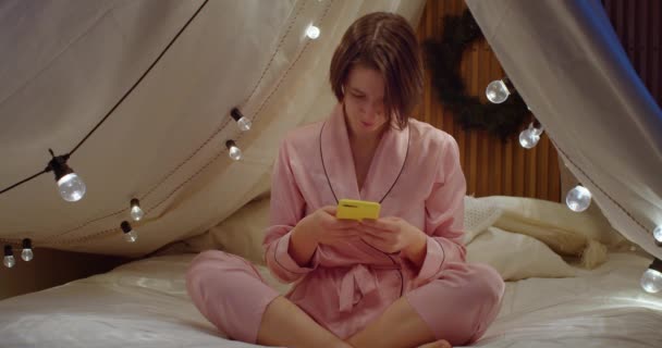 Mladá žena sedící se zkříženýma nohama a smartphonem uvnitř v útulné posteli. Atraktivní dívka rolování zpravodajství na mobilním telefonu před spaním. Dámské módní oblečení. Koncept aplikačních technologií — Stock video