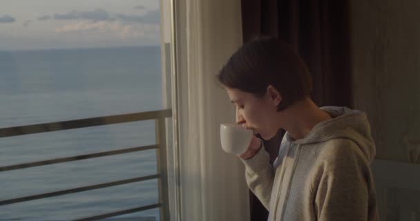 Side portrait of young girl drinking tea looking outside and smiling. Attractive woman with cup in hand enjoying beautiful view and dreaming. Peaceful evening at home — 비디오