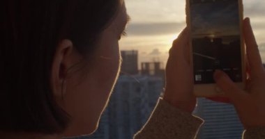 Akıllı telefon cihazıyla gün batımının fotoğraflarını çeken genç bir kadın. Güzel şehir manzarasından hoşlanan ve akşamları binaları ve mimarileri fotoğraflayan bir kız. Cep telefonu fotoğrafçılığı 