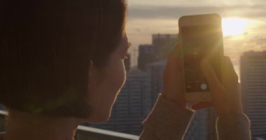 Akıllı telefon cihazıyla gün batımının fotoğraflarını çeken genç bir kadın. Güzel şehir manzarasından hoşlanan ve akşamları binaları ve mimarileri fotoğraflayan bir kız. Cep telefonu fotoğrafçılığı 