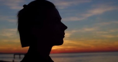 Sahilde akşam ışığında hayal kuran arka ışıklandırmalı kızın yakın plan portresi. Günbatımında kadın profili silueti renkli gökyüzü arka planında keyif çatıyor. Deniz kıyısında dinlenme ve meditasyon.