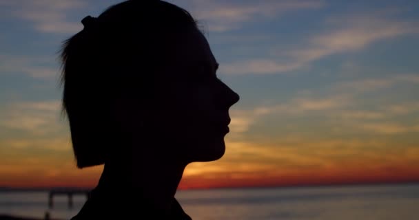 Közelkép oldalsó portré a háttérvilágítású lányról, aki a tengerparton álmodik természetes esti fényben. Női profil sziluett naplemente színes ég háttér élvezi. Pihenés és meditáció a tengerparton a tenger közelében — Stock videók
