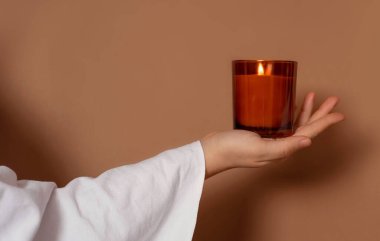 Covered with white cloth hand holding a candle on a brown background. Marketing banner for candle shop. Aromatherapy poster. Boho atmosphere for scent products. AD banner for spa. High quality photo