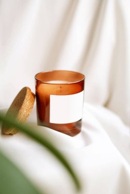 Boş etiketli cam kavanozdaki mumu kapat. Bulanık yeşil bitki, beyaz ipek arka plan. Ön manzara. El yapımı ekolojik ve vejetaryen soya mumları. Model yap, mesaj için yer aç. Yüksek kalite fotoğraf