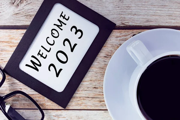 Welcome 2023 text on chalkboard frame and coffee on wooden desk. New year concept.