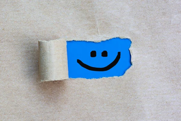 Happy smiley face on torn hole in the sheet of brown paper on blue background.