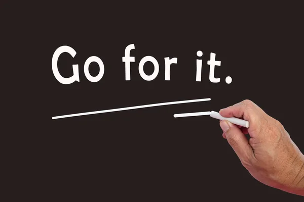 Male hand writes in white chalk pencil the word Go For It on a black background. Encouragement phrases and business concept.