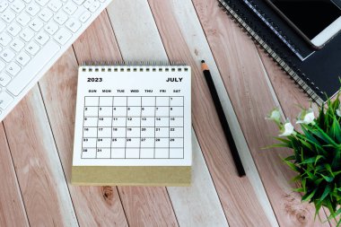 White July 2023 calendar on wooden desk with keyboard, note books, pencil, potted plant and smartphone.