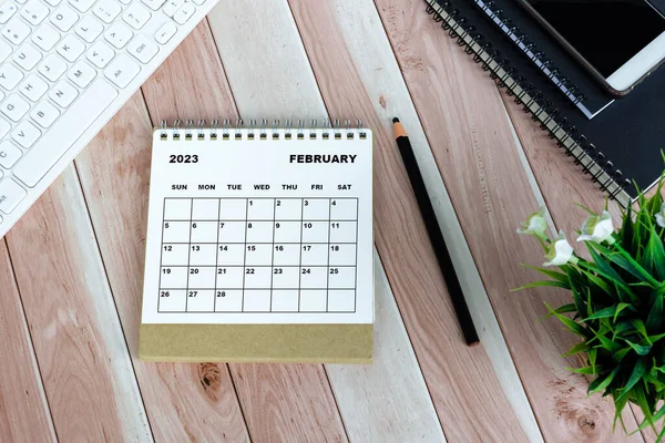 White February 2023 calendar on wooden desk with keyboard, note books, pencil, potted plant and smartphone.