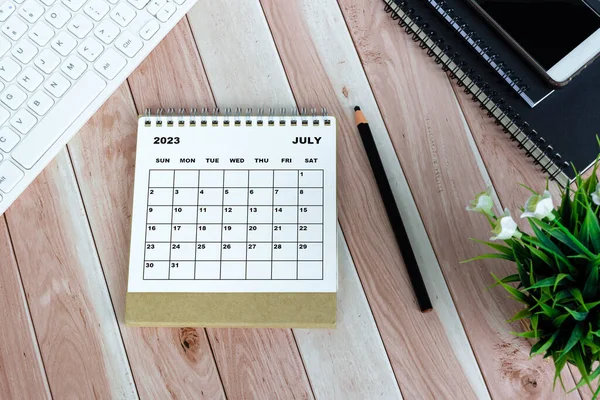 White July 2023 calendar on wooden desk with keyboard, note books, pencil, potted plant and smartphone.